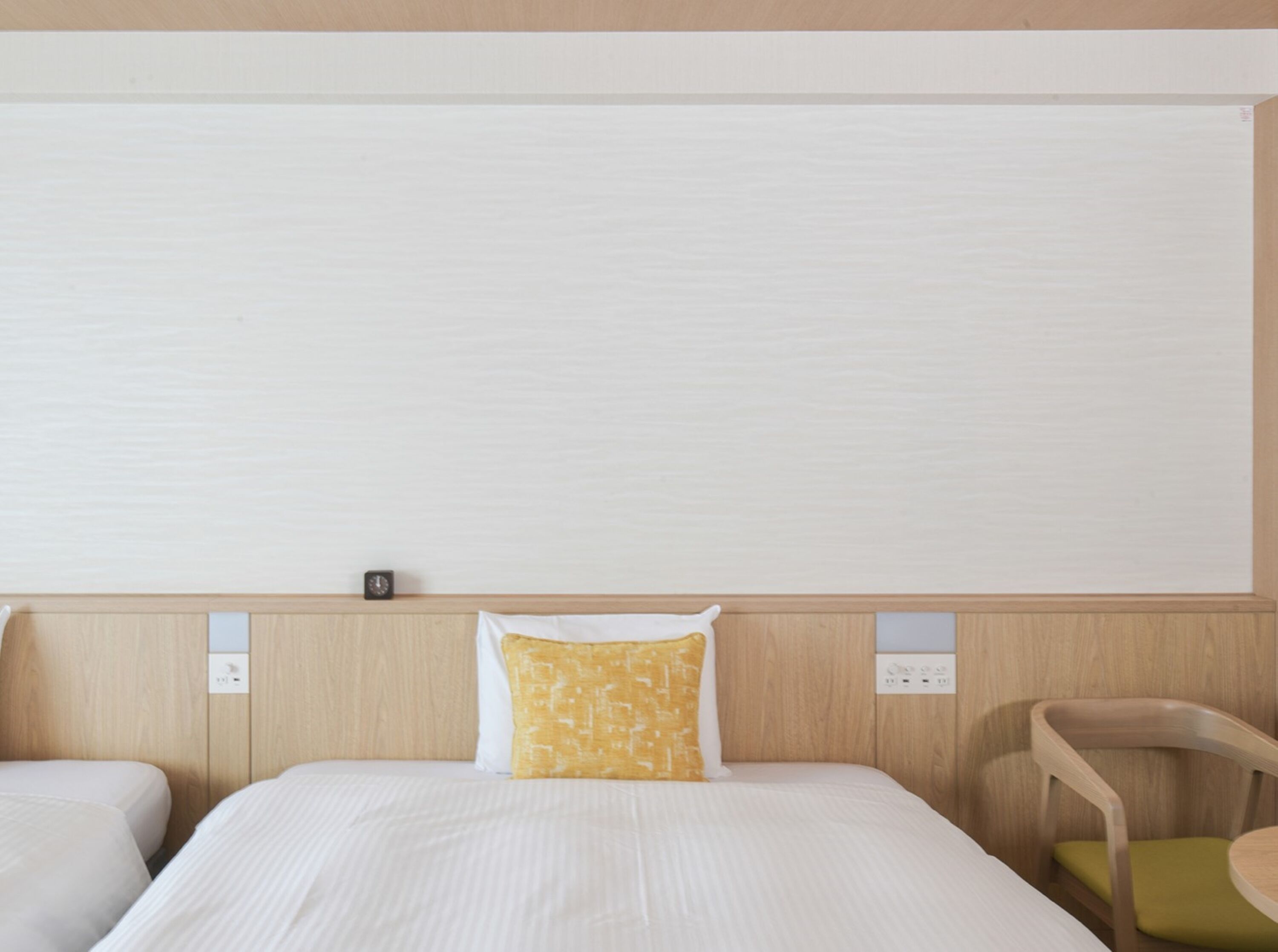 Hotellihuone, jossa on sänky, yöpöytä, tuoli ja seinälle asennettu ohjauspaneeli.