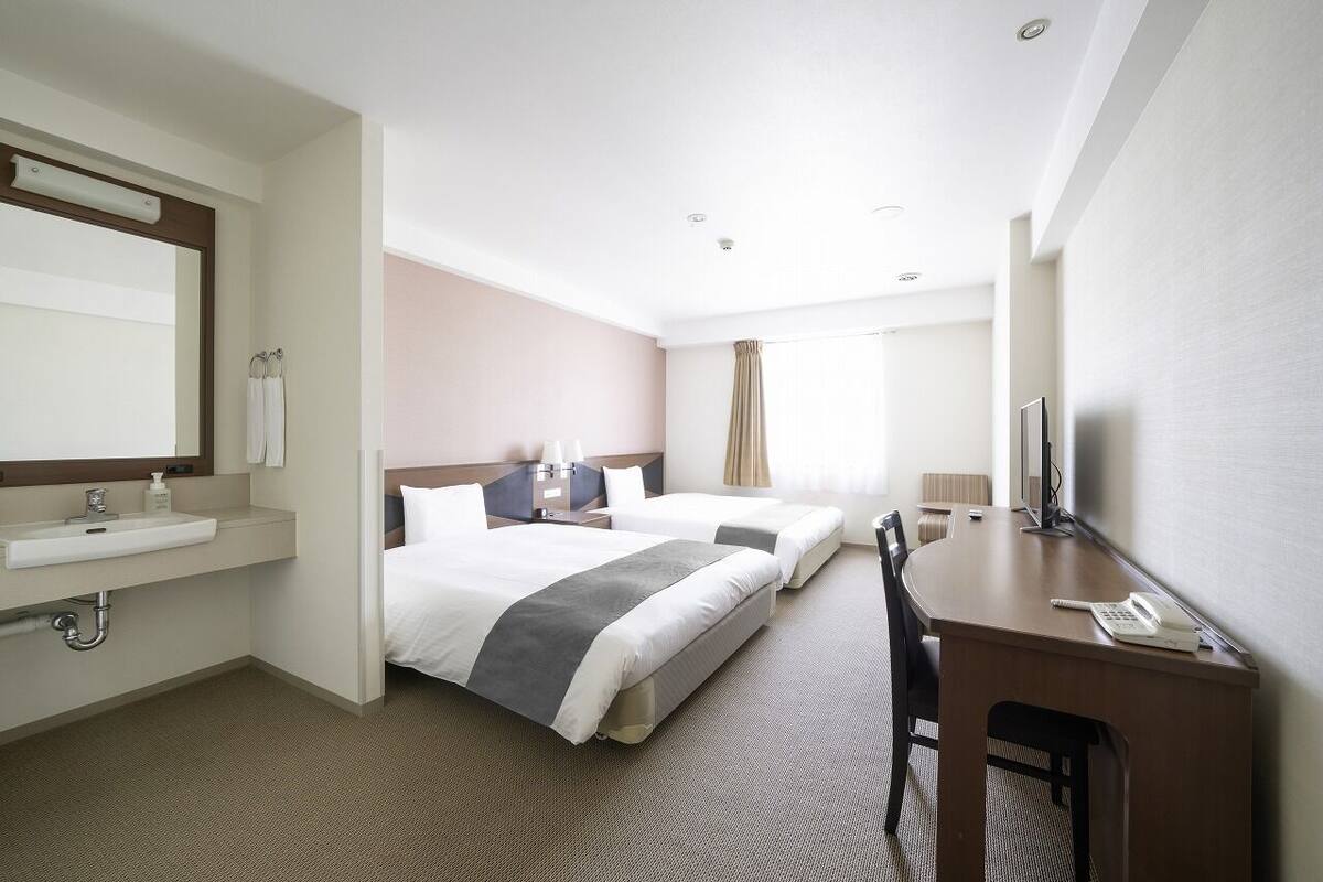 Ett hotellrum med två sängar, ett skrivbord, en stol, en TV, ett badrum med handfat och spegel samt ett fönster med gardiner.