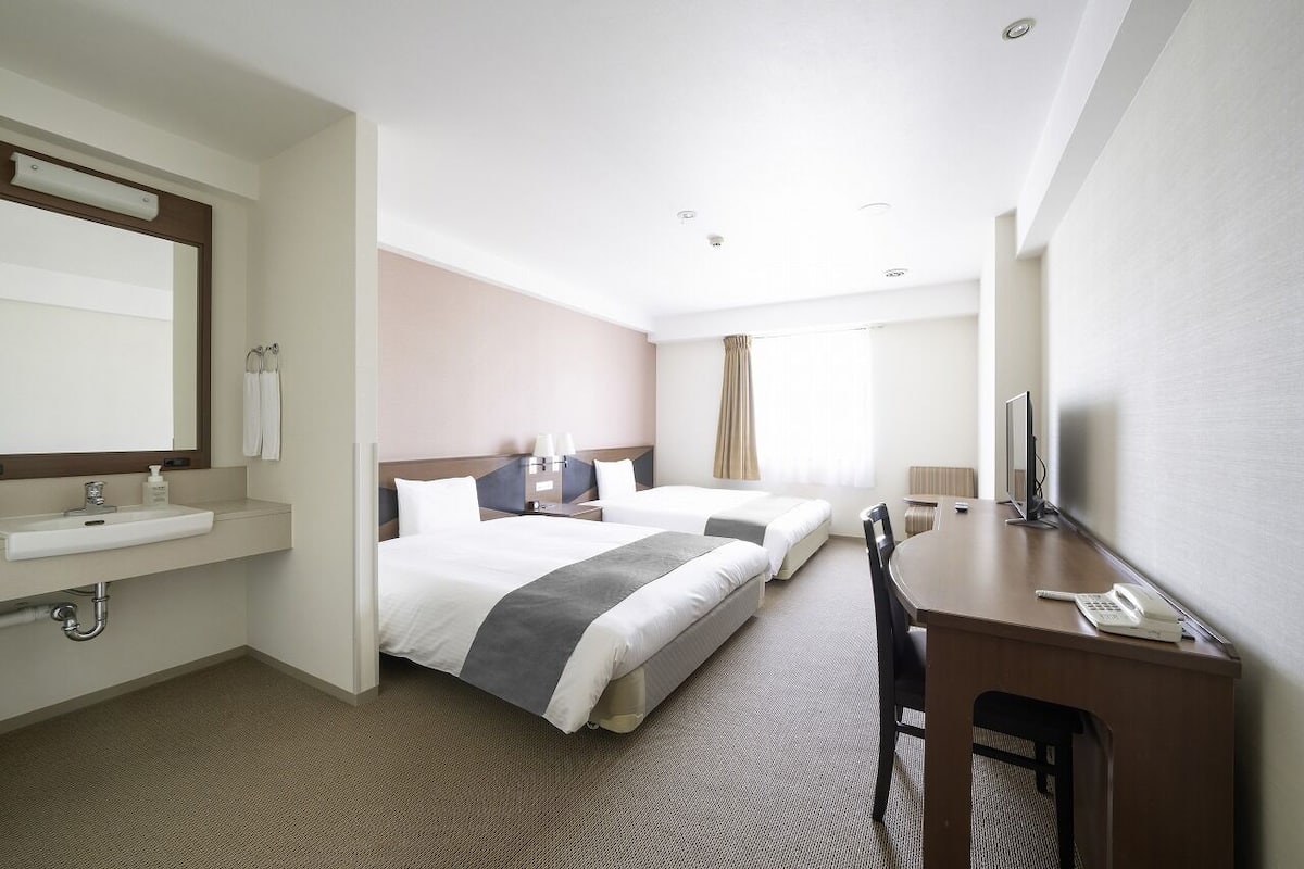 A hotel room with two beds, a desk, a chair, a TV, a bathroom with a sink and mirror, and a window with curtains.