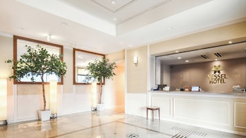 A hotel reception area with a modern design, including a reception desk, two potted plants, and a large mirror.