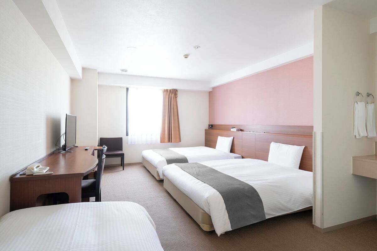 Ett hotellrum med två sängar, ett skrivbord med en dator, en stol, ett litet bord, ett fönster med gardiner och ett badrum med en spegel och en handduksställning.