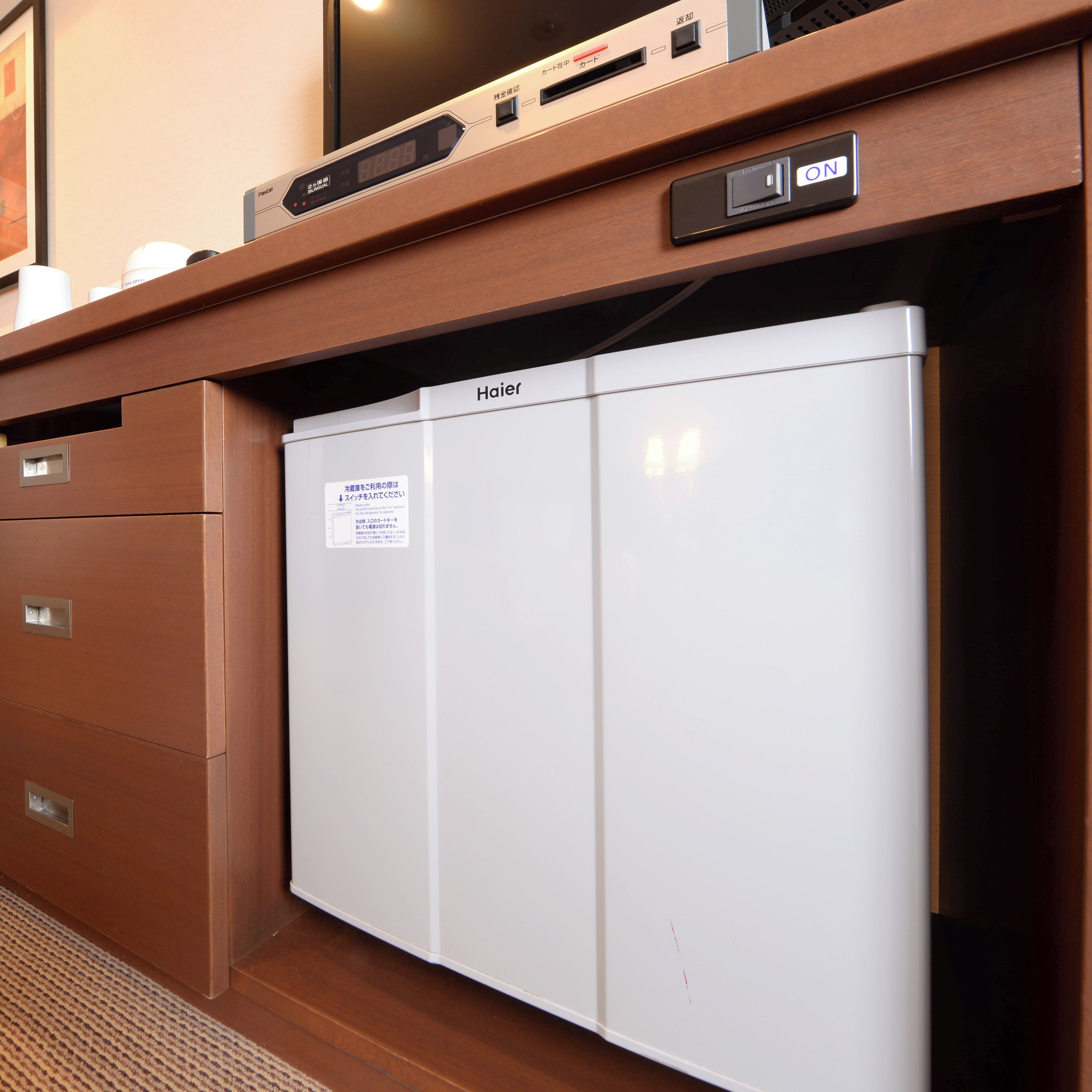 A compact, white refrigerator with the brand name 'Haier' on the door, situated in a wooden cabinet with a digital clock above it.