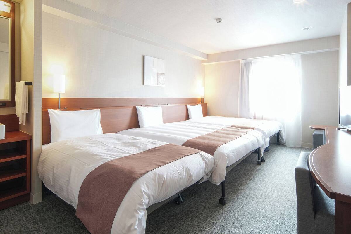 Ett hotellrum med två sängar, ett skrivbord, en spegel och ett fönster med gardiner.