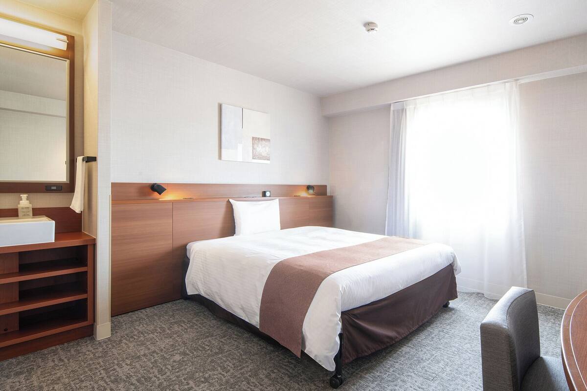 Ett hotellrum med en säng, ett skrivbord, en TV, ett badrum med spegel och handfat samt ett fönster med gardiner.
