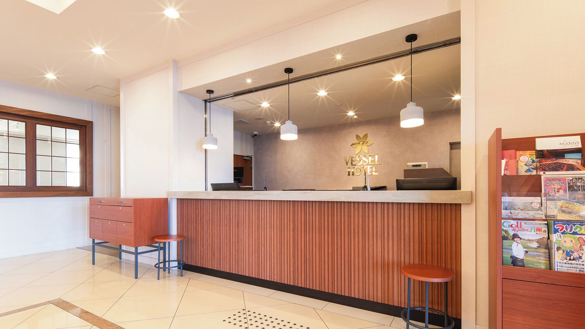 A hotel reception area with a wooden reception desk, two bar stools, a bulletin board with magazines, and a large window with blinds.
