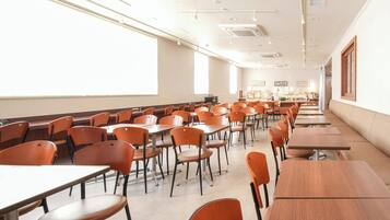 A classroom with rows of desks and chairs, a projector screen, and a kitchenette area.