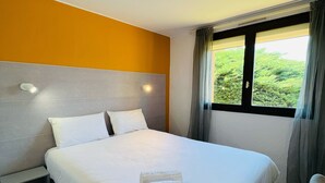 A hotel room with a large bed, two bedside tables, a window with curtains, and a view of greenery outside. - Hôtel Le Forestia (Pannes)