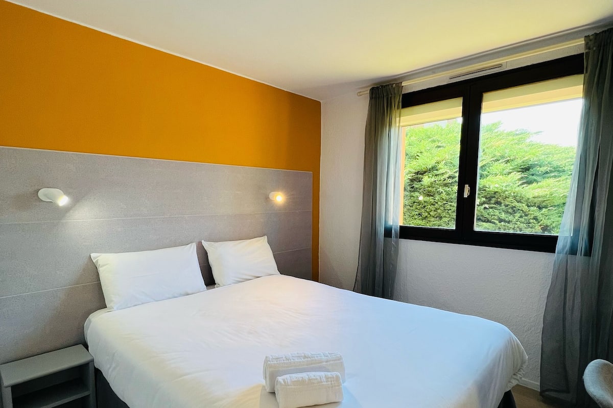 A hotel room with a large bed, two bedside tables, a window with curtains, and a view of greenery outside.