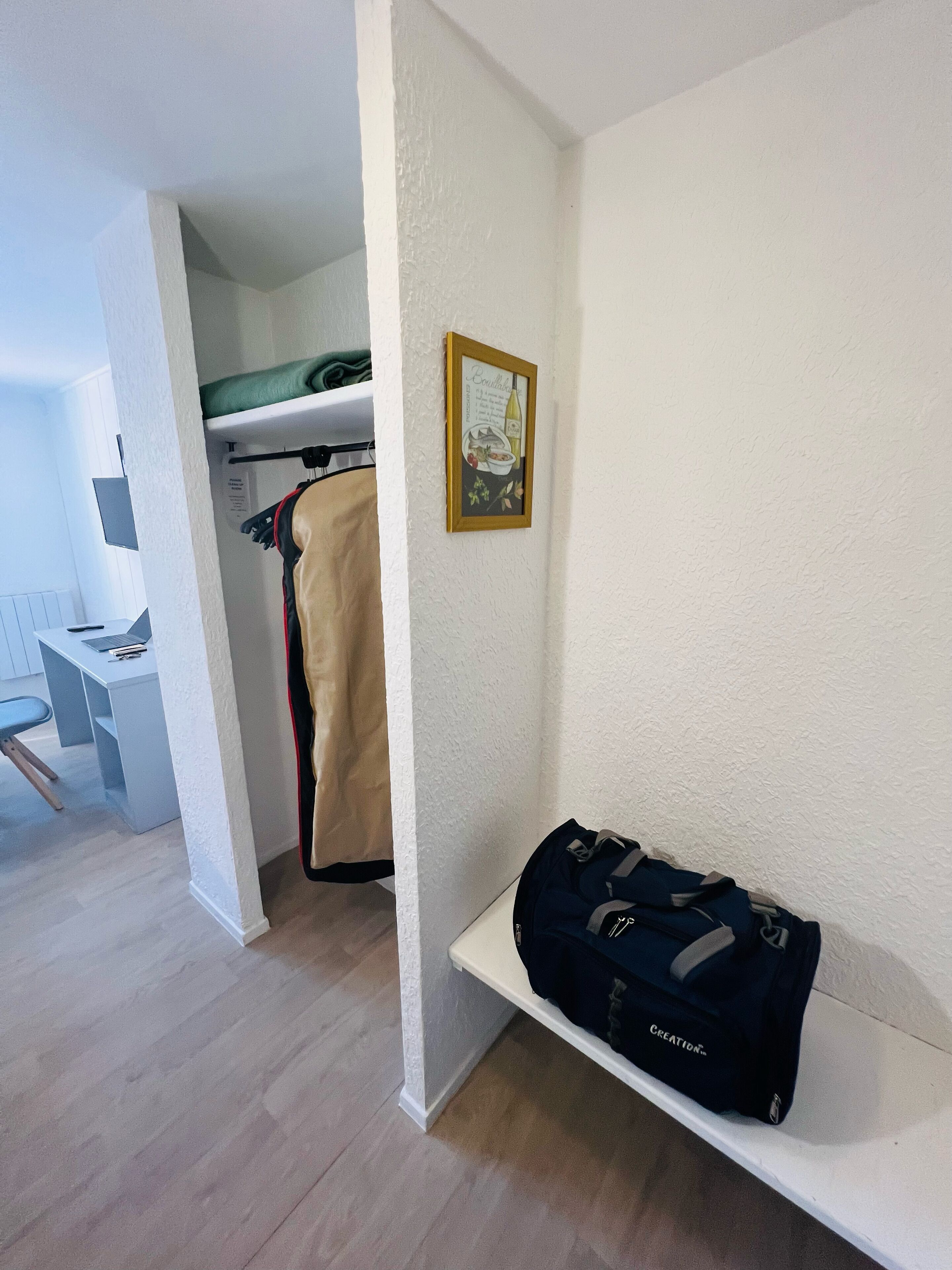 Une chambre d’hôtel avec un lit, un bureau et une chaise, un porte-manteau et une étagère avec un sac.