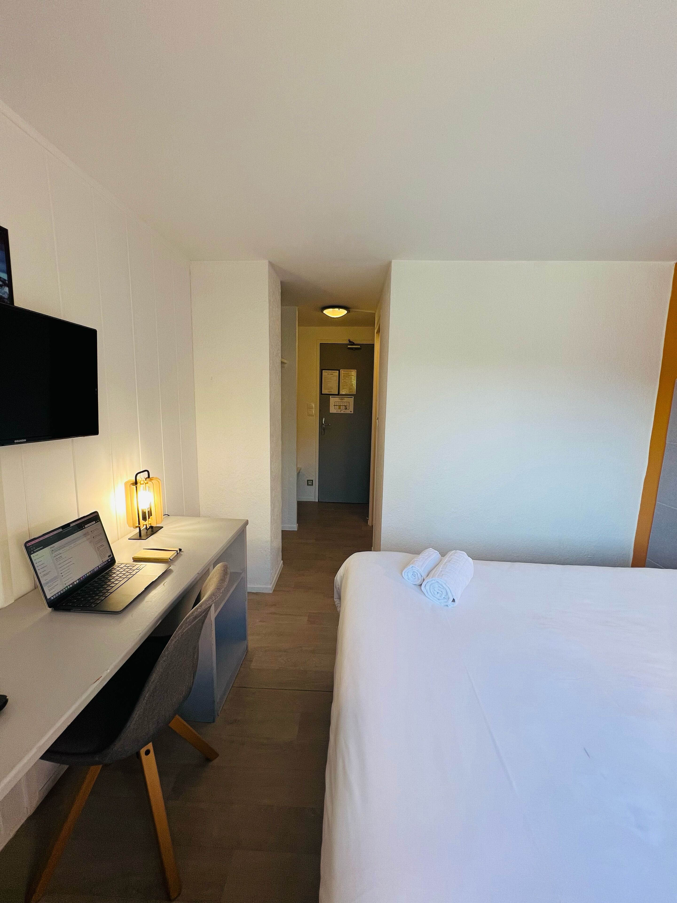 A hotel room with a bed, a desk with a laptop, a lamp, and a TV mounted on the wall.