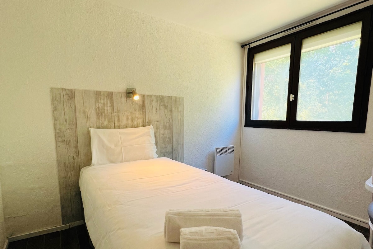 A hotel room with a large bed, a wooden headboard, a window with blinds, and a bedside table.