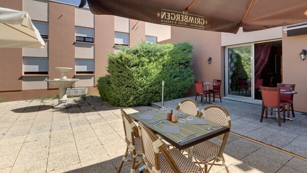 An outdoor patio area with a table and chairs, a large umbrella, and a building with a sign reading "GRIMBERGEN". - Hôtel Le Forestia (Pannes)