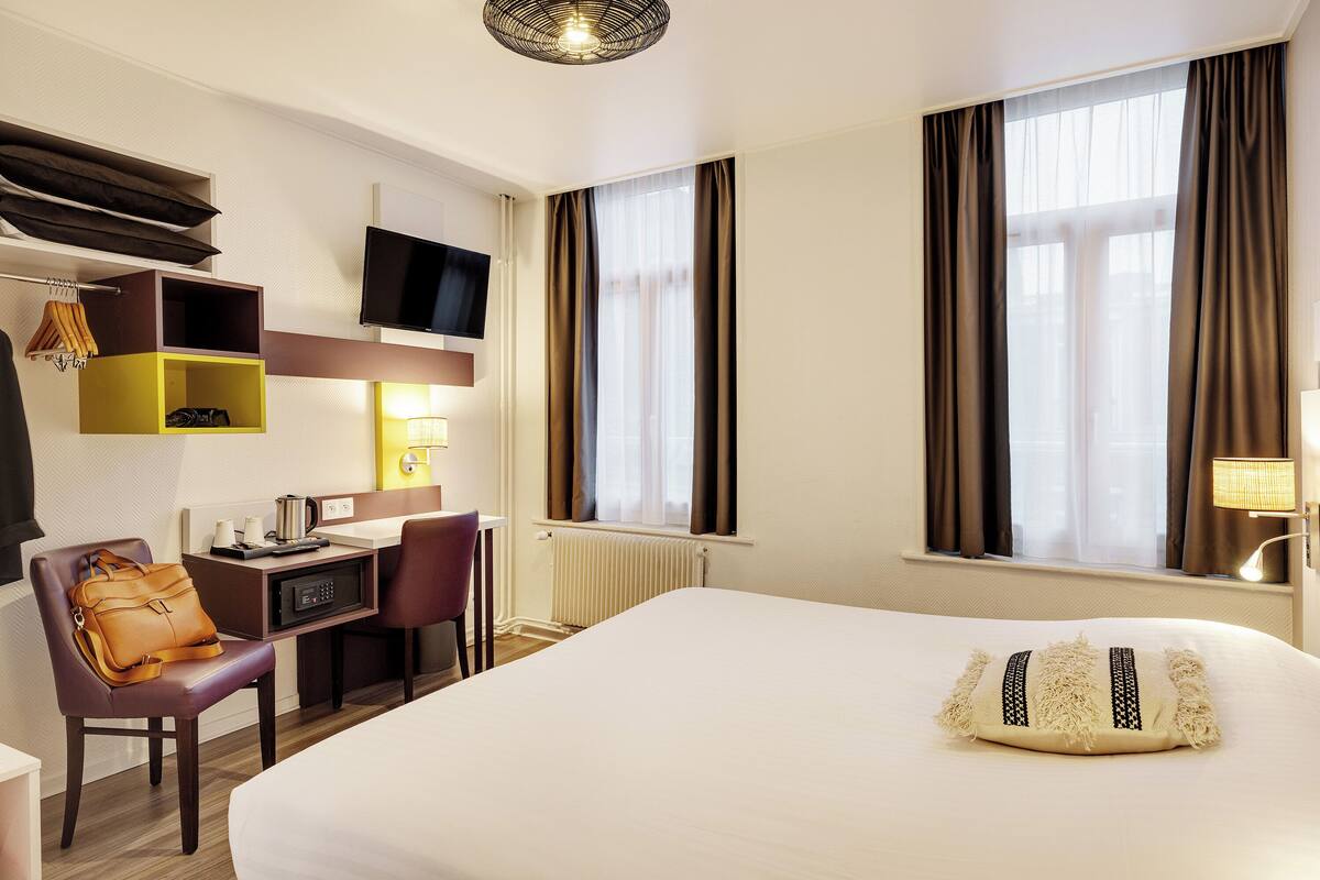 A hotel room with a bed, a desk, a chair, a TV, and a window with curtains.