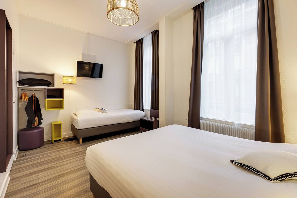 A hotel room with a large bed, a bedside table, a lamp, a TV mounted on the wall, and a window with curtains.