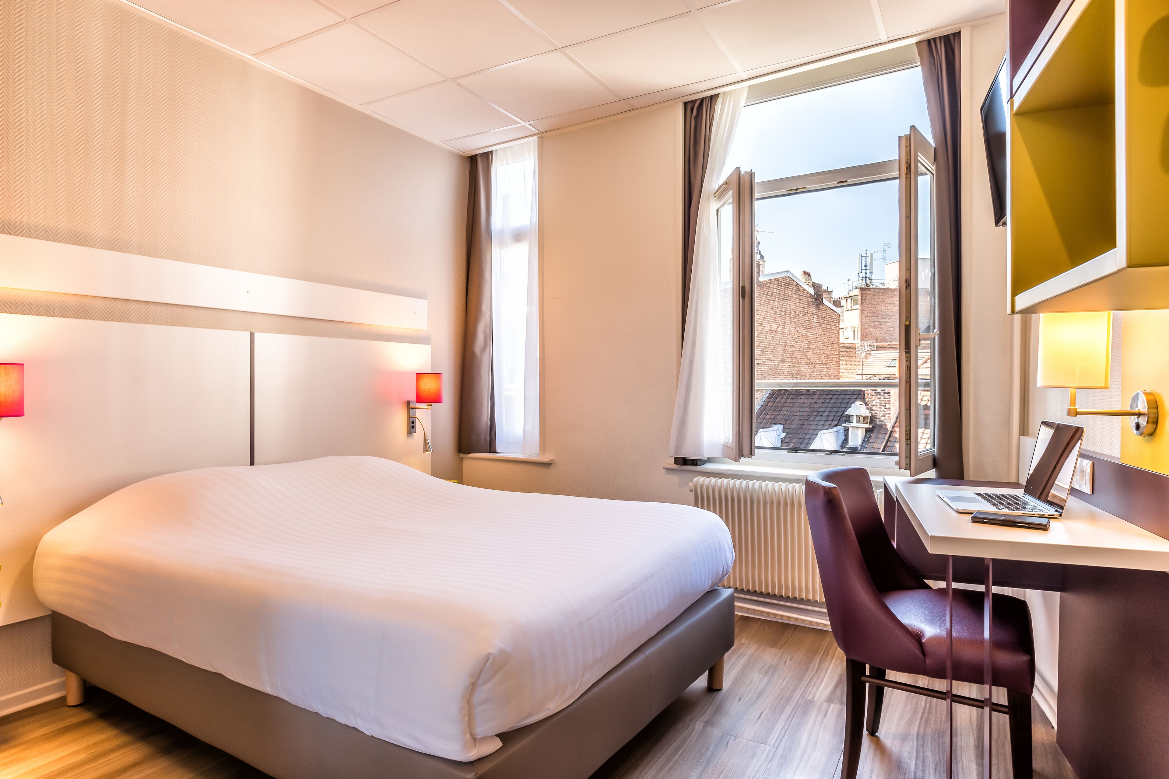 Habitación de hotel con cama, escritorio con computadora portátil, una silla, ventana con cortinas y vista a edificios.