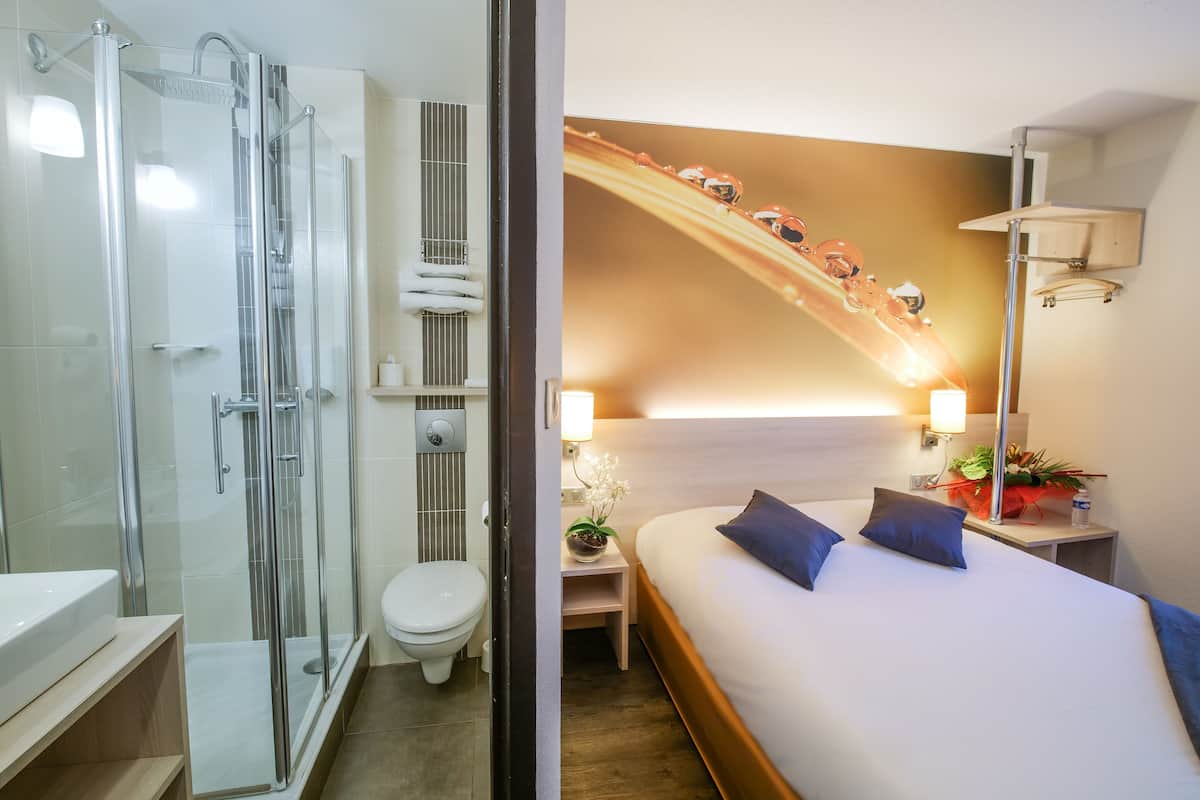 A hotel room with a glass-enclosed shower, a toilet, a bed with two pillows, a nightstand with a lamp, and a wall-mounted shelf with a plant.