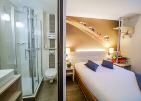 Habitación de hotel con ducha acristalada, inodoro, cama con dos almohadas, mesita de noche con lámpara y estante montado en la pared con una planta.