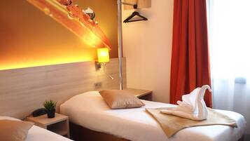 A hotel room with two beds, a bedside table with a lamp, a wall-mounted light, and a window with curtains.