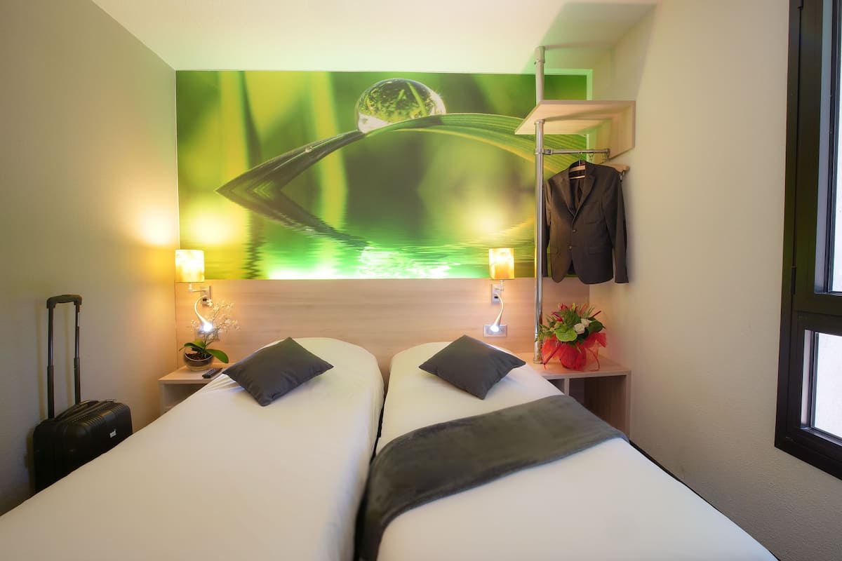 A hotel room with two beds, a nightstand with a lamp, a wall-mounted shelf with a jacket, and a window.