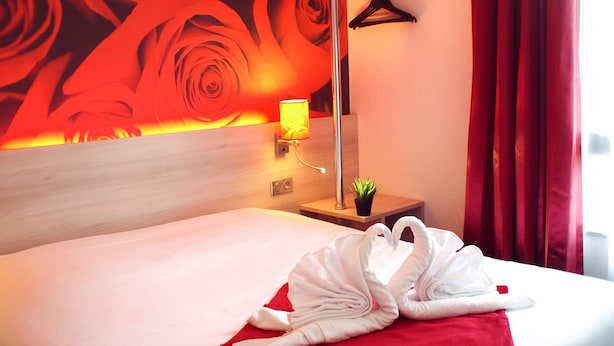 A hotel room with a bed, a red pillow, a towel, a nightstand, a lamp, a small plant, and a red curtain.