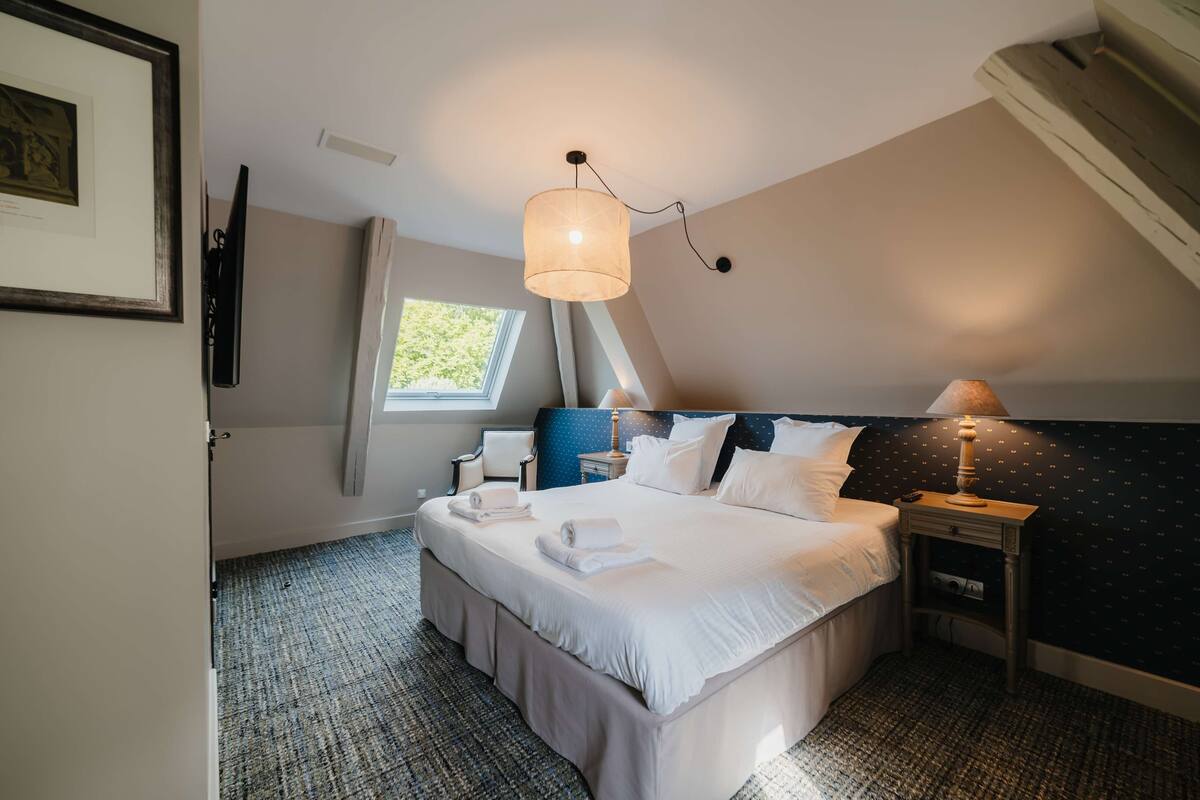 Un lit bien fait, avec du linge de lit blanc, une lampe de chevet et une petite table sur laquelle se trouve une autre lampe.