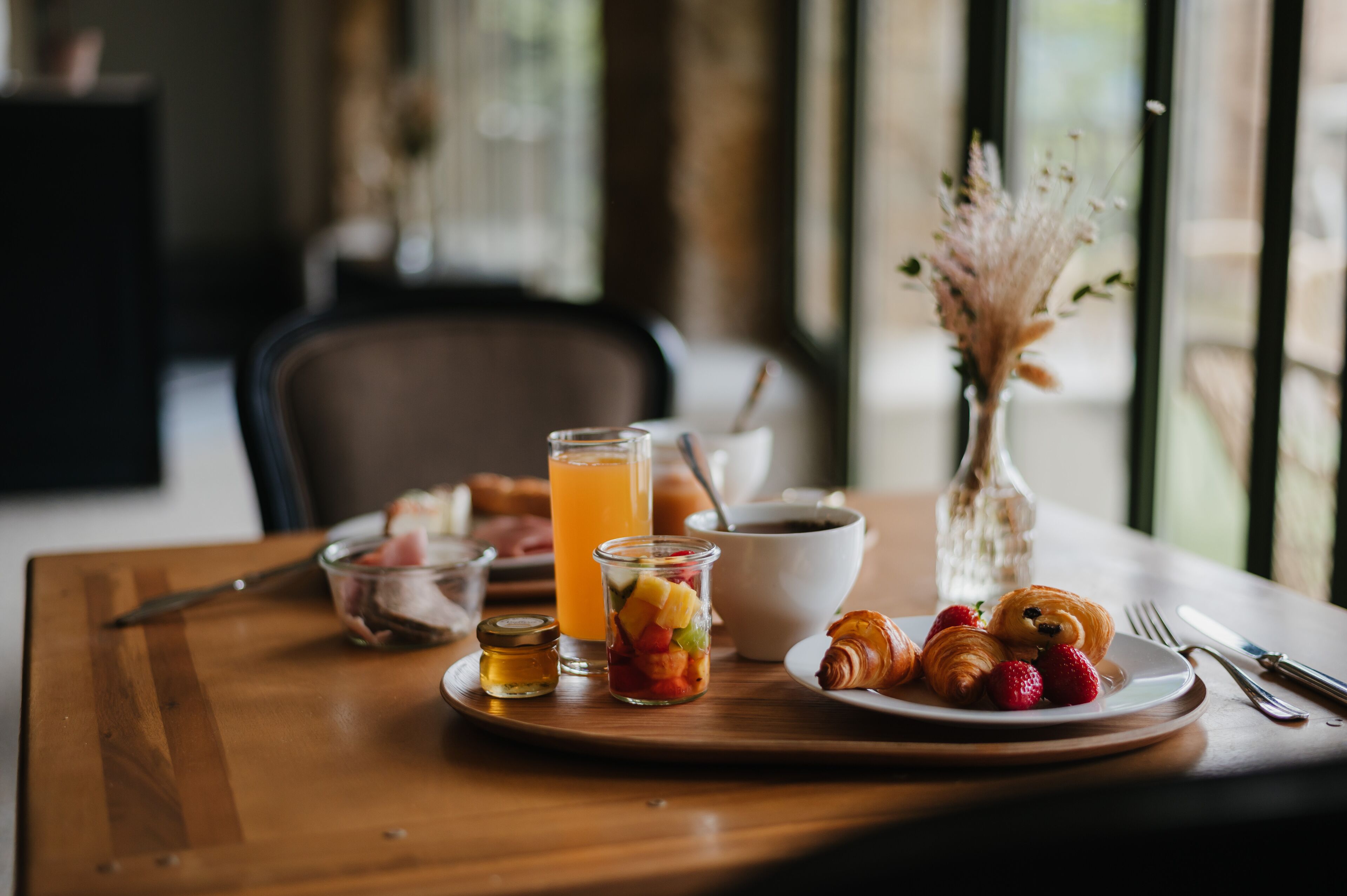 Una mesa de desayuno con cruasanes, fresas, un vaso de jugo de naranja, un tazón de cereal y un frasco pequeño de mermelada.