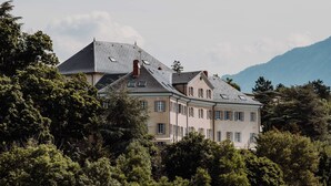 A large, multi-story building with a symmetrical facade, surrounded by dense greenery and mountains in the background. - Hotel La Robeyere, BW Signature Collection (Embrun)
