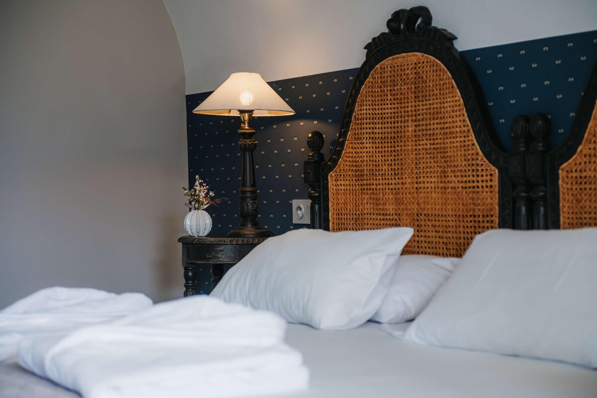 Un lit bien fait, avec du linge de lit blanc, une table de chevet en bois foncé surmontée d’une lampe, et un mur décoratif orné d’un motif géométrique.