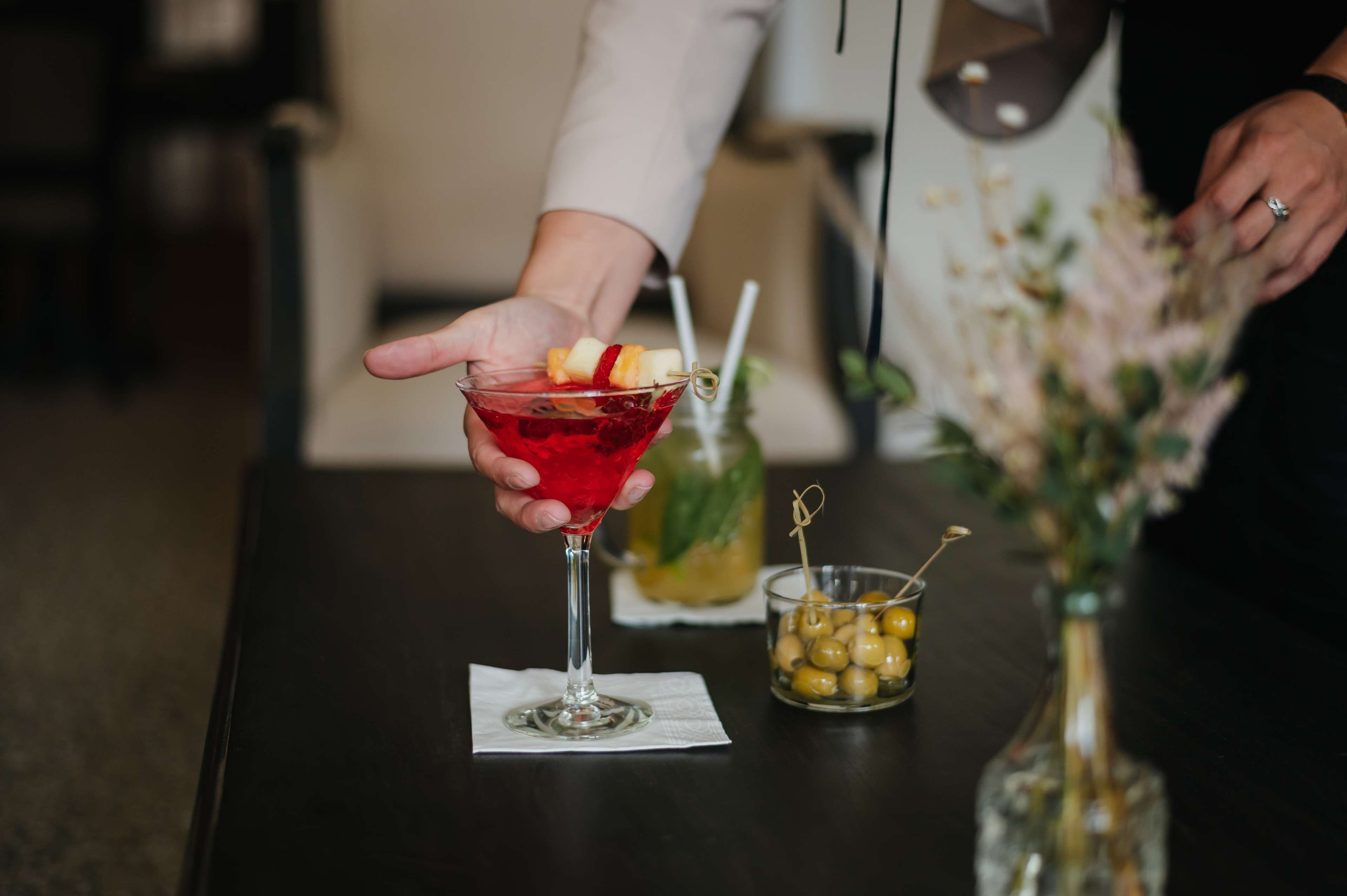 A hand holding a cocktail with fruit garnish, a bottle of olive oil, and a small bowl of olives on a table.