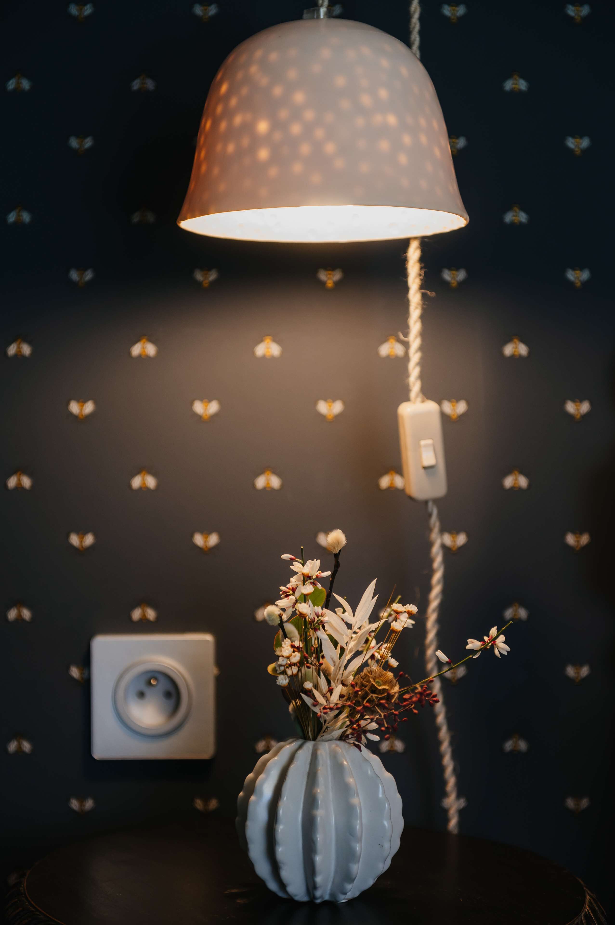 A lamp with a textured shade, a vase with flowers, and a power outlet on a wall with a heart-patterned wallpaper.