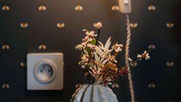 Una lámpara con pantalla texturizada, un jarrón con flores y un enchufe en una pared con papel tapiz estampado con corazones.