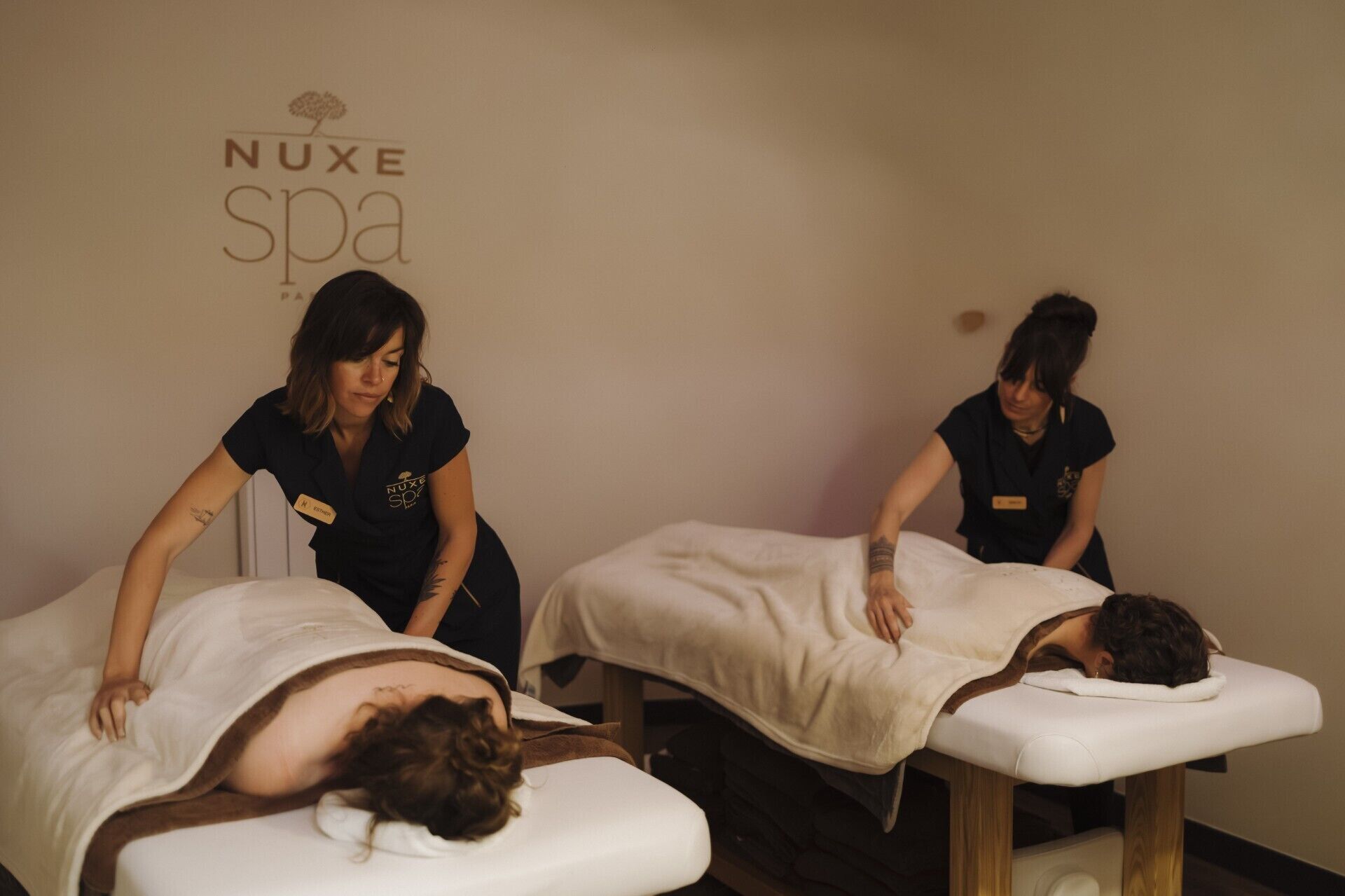 A spa room with two massage tables, one occupied by a client and two staff members attending to clients.