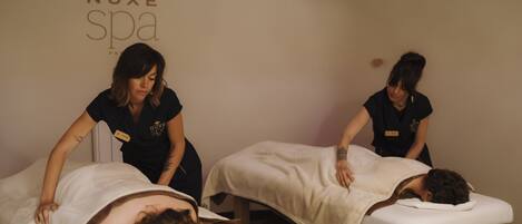 A spa room with two massage tables, one occupied by a client and two staff members attending to clients.