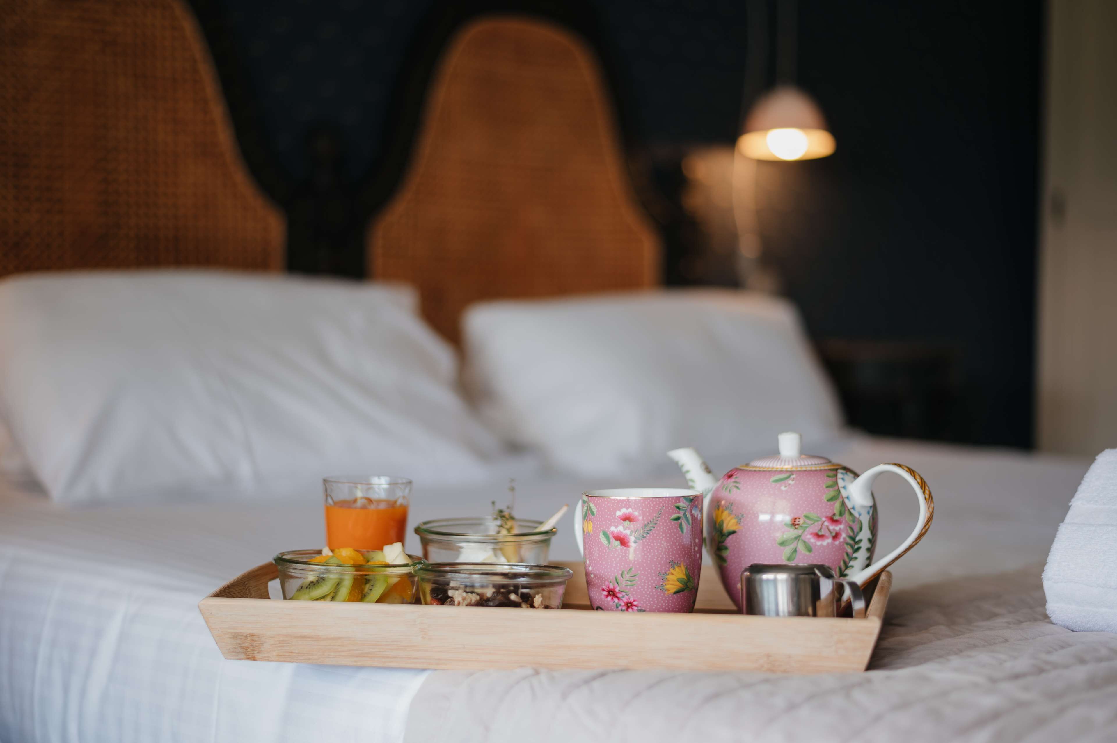 A wooden tray with breakfast items on a bed, including a glass of orange juice, a teapot, and a cup with floral design.