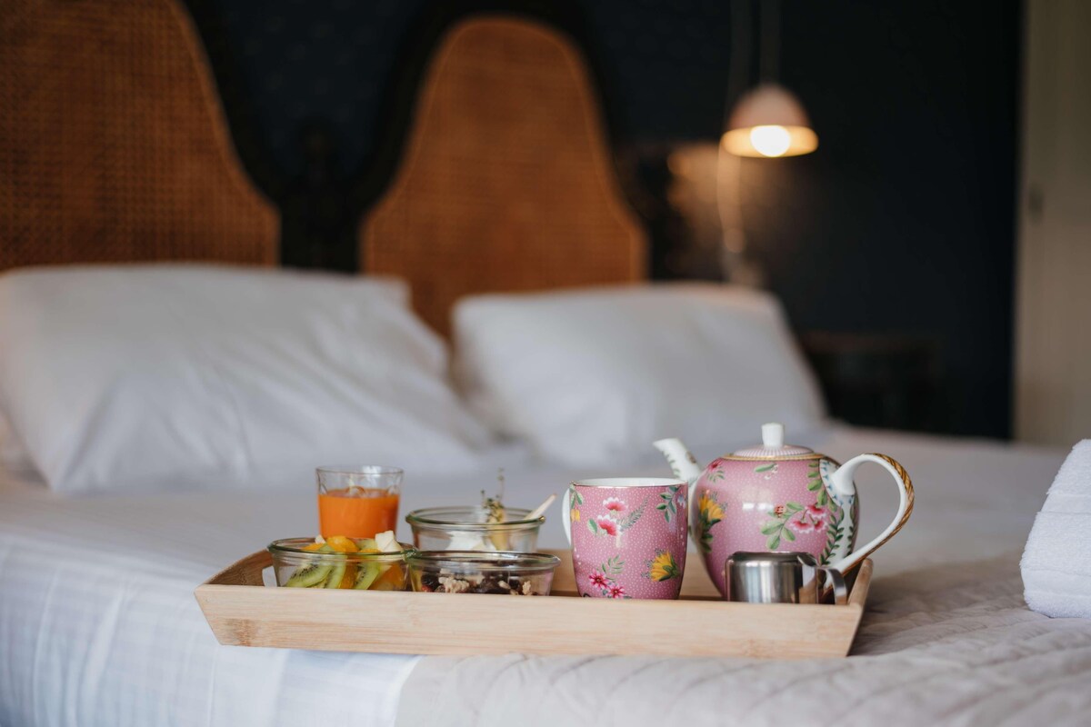 A neatly arranged breakfast tray on a bed with a floral teapot, a glass of orange juice, and a jar of mixed nuts.