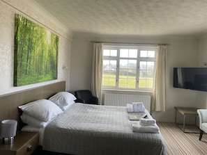 A hotel room with a large bed, a flat-screen TV, a window with curtains, and a painting of a forest.