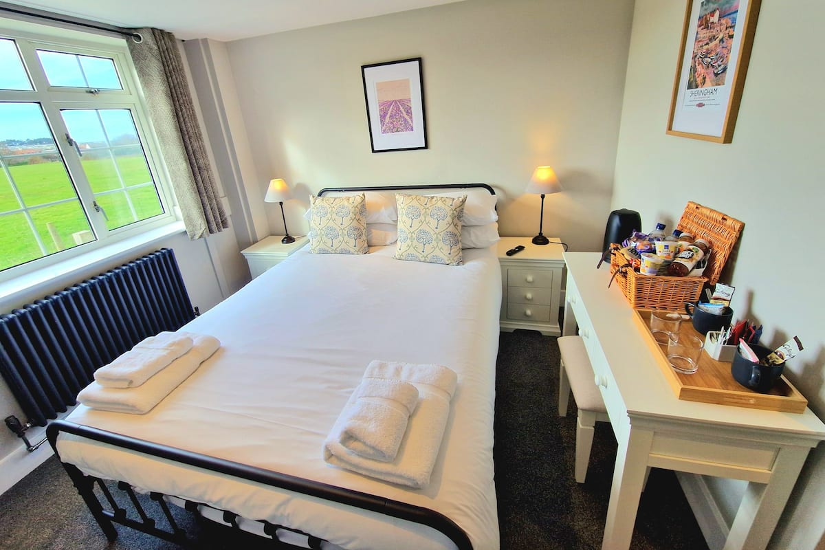 A neatly made bed with white linens and towels, a bedside table with a lamp, a framed picture on the wall, and a view of a green field through the window.