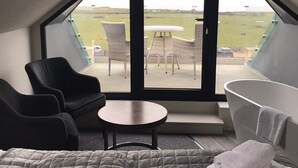 A bedroom with a large bed, a sitting area with chairs, a small table, a bathtub, and a view of the outdoors. - Dormy House Hotel (Cromer)