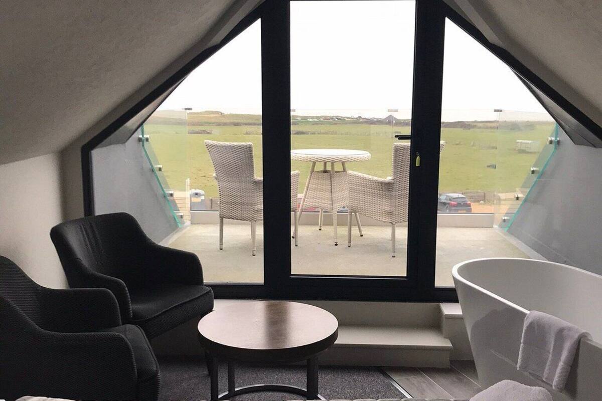A bedroom with a large bed, a sitting area with chairs, a small table, a bathtub, and a view of the outdoors.