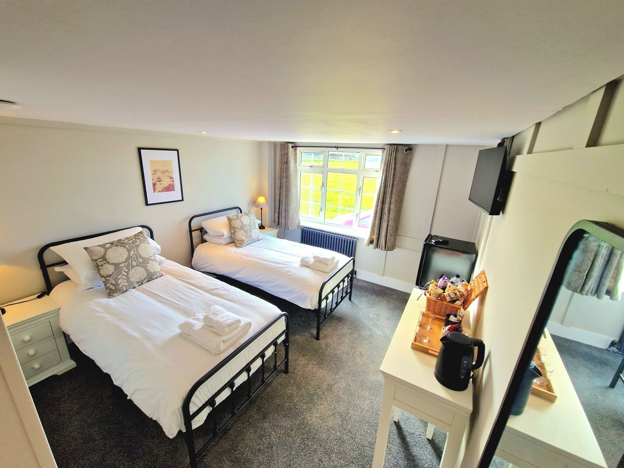 A hotel room with two beds, a desk with a kettle, a TV, and a window with curtains.
