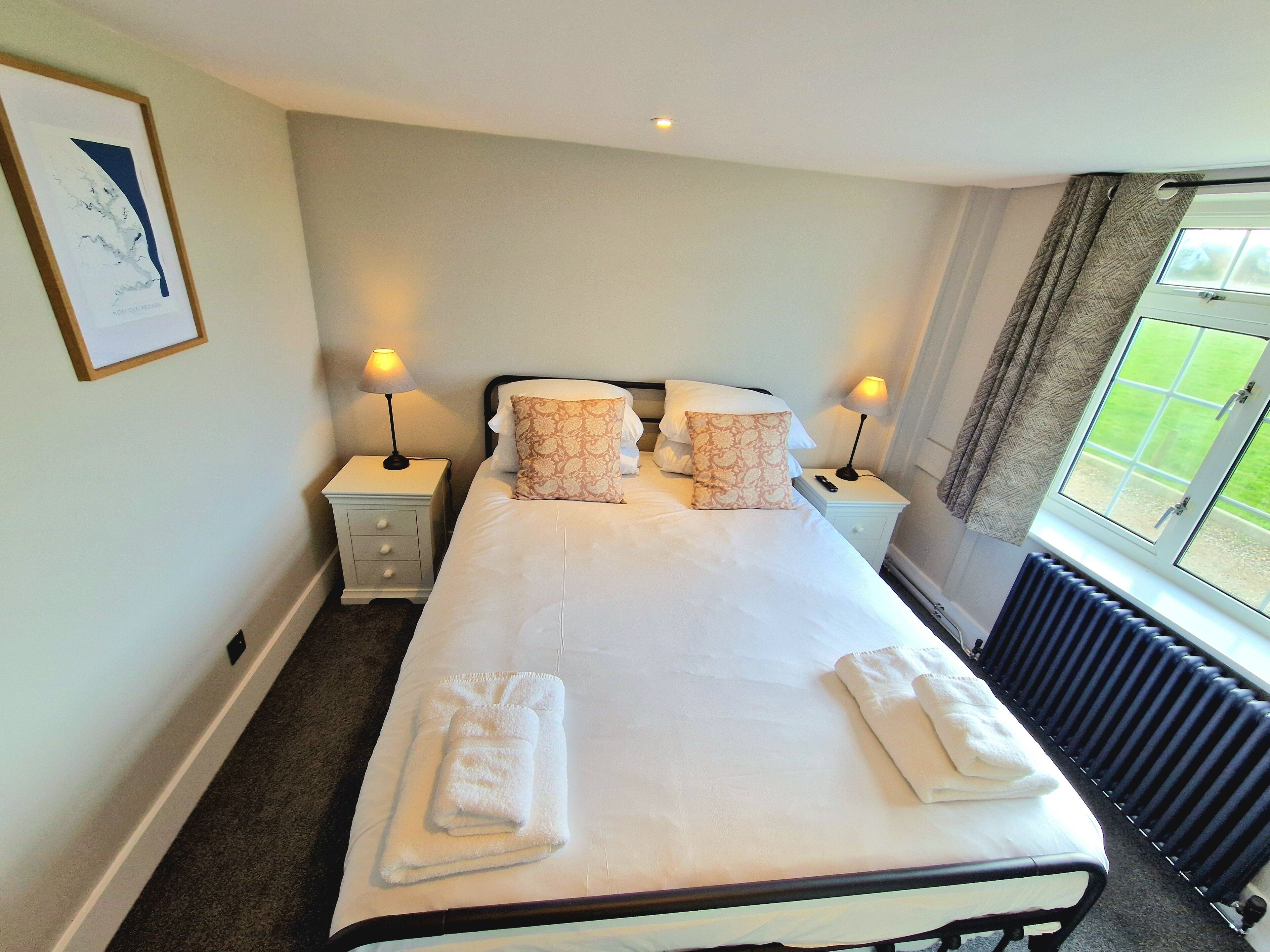 A neatly made bed with white linens and two decorative pillows, two bedside tables with lamps, a framed picture on the wall, and a window with curtains.
