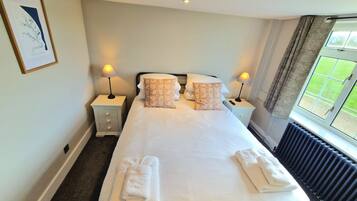A neatly made bed with white linens and two decorative pillows, two bedside tables with lamps, a framed picture on the wall, and a window with curtains.