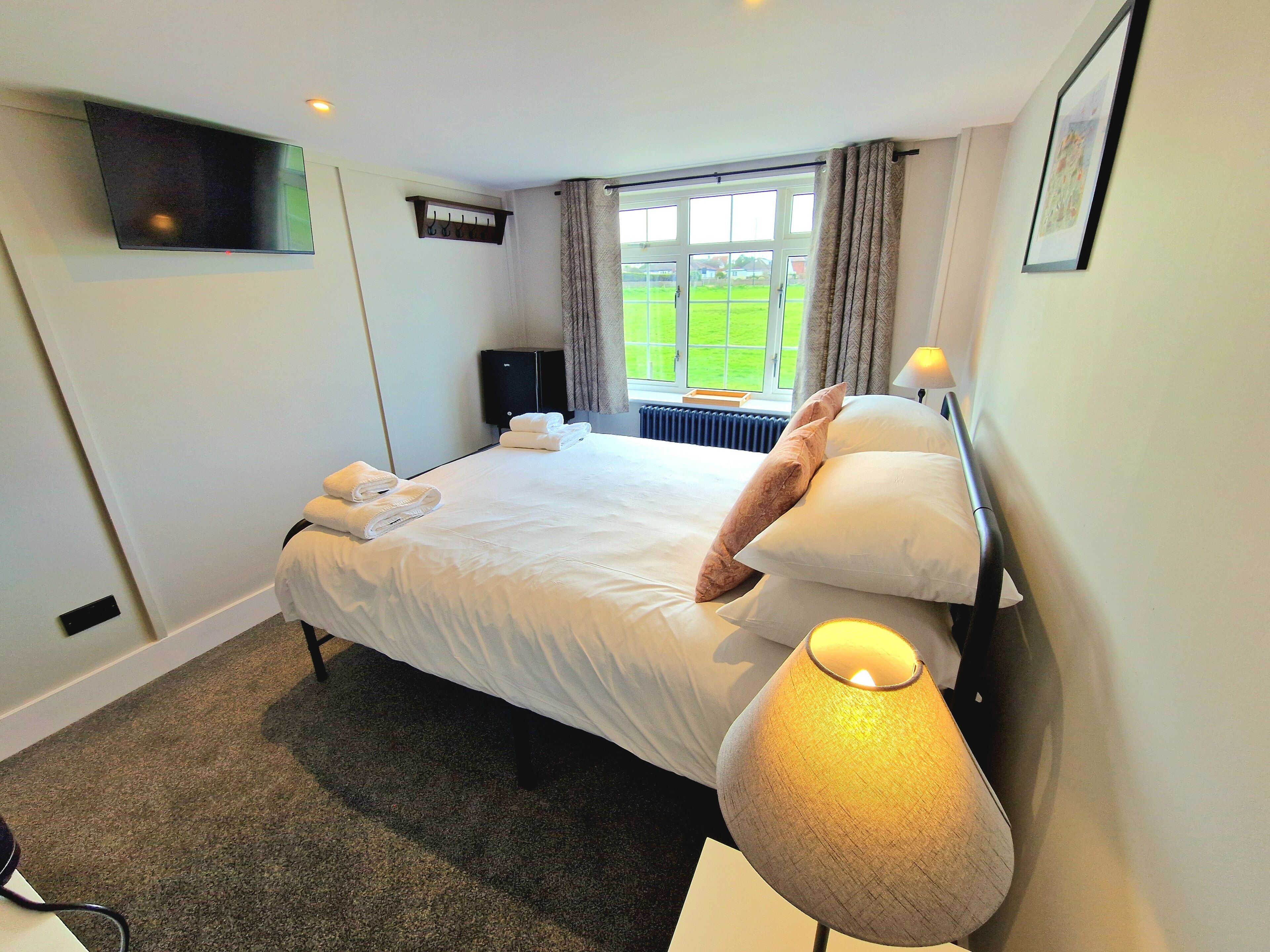 A hotel room with a bed, a flat-screen TV mounted on the wall, a window with curtains, a radiator, and a bedside lamp.