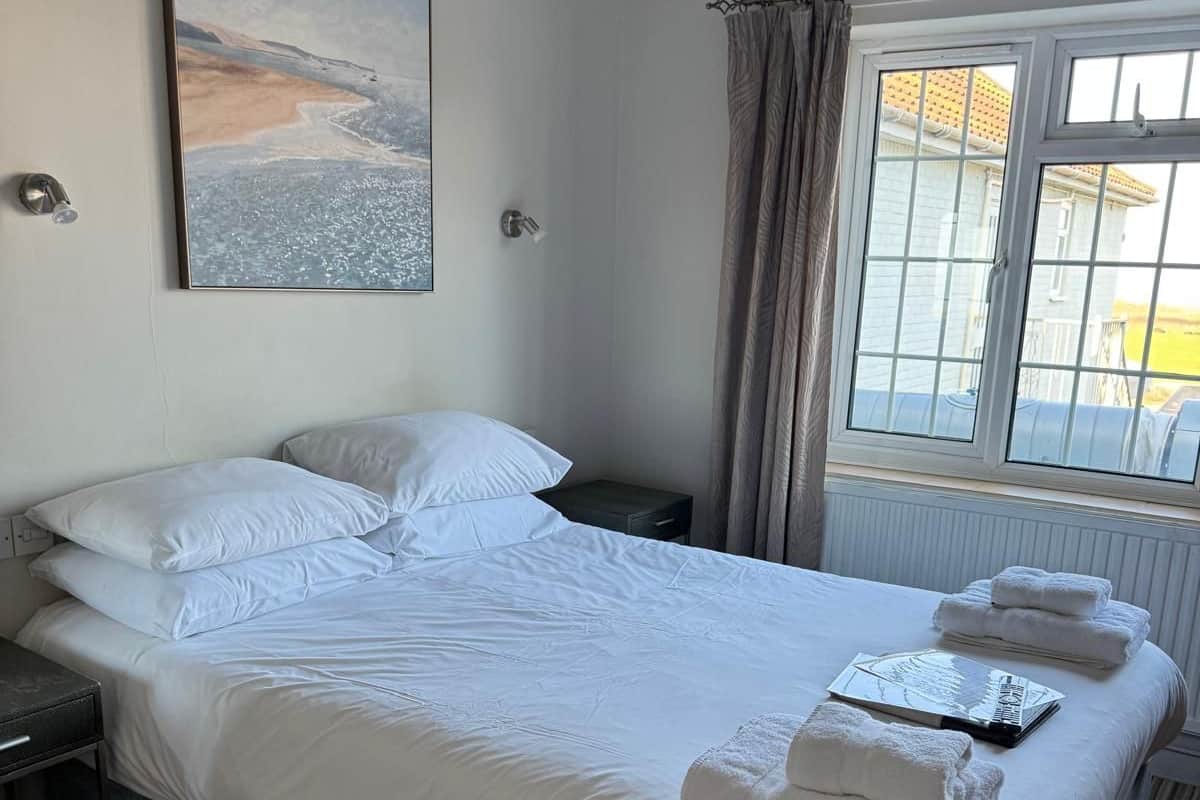 A neatly made bed with white linens and towels, a bedside table with a lamp, a window with curtains, and a painting on the wall.