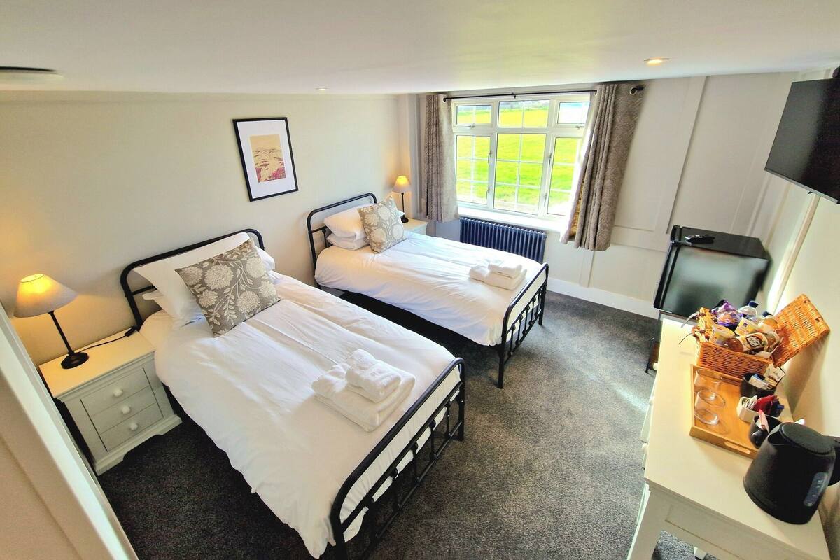 A hotel room with two beds, a desk with a kettle, a television, and a window with curtains.