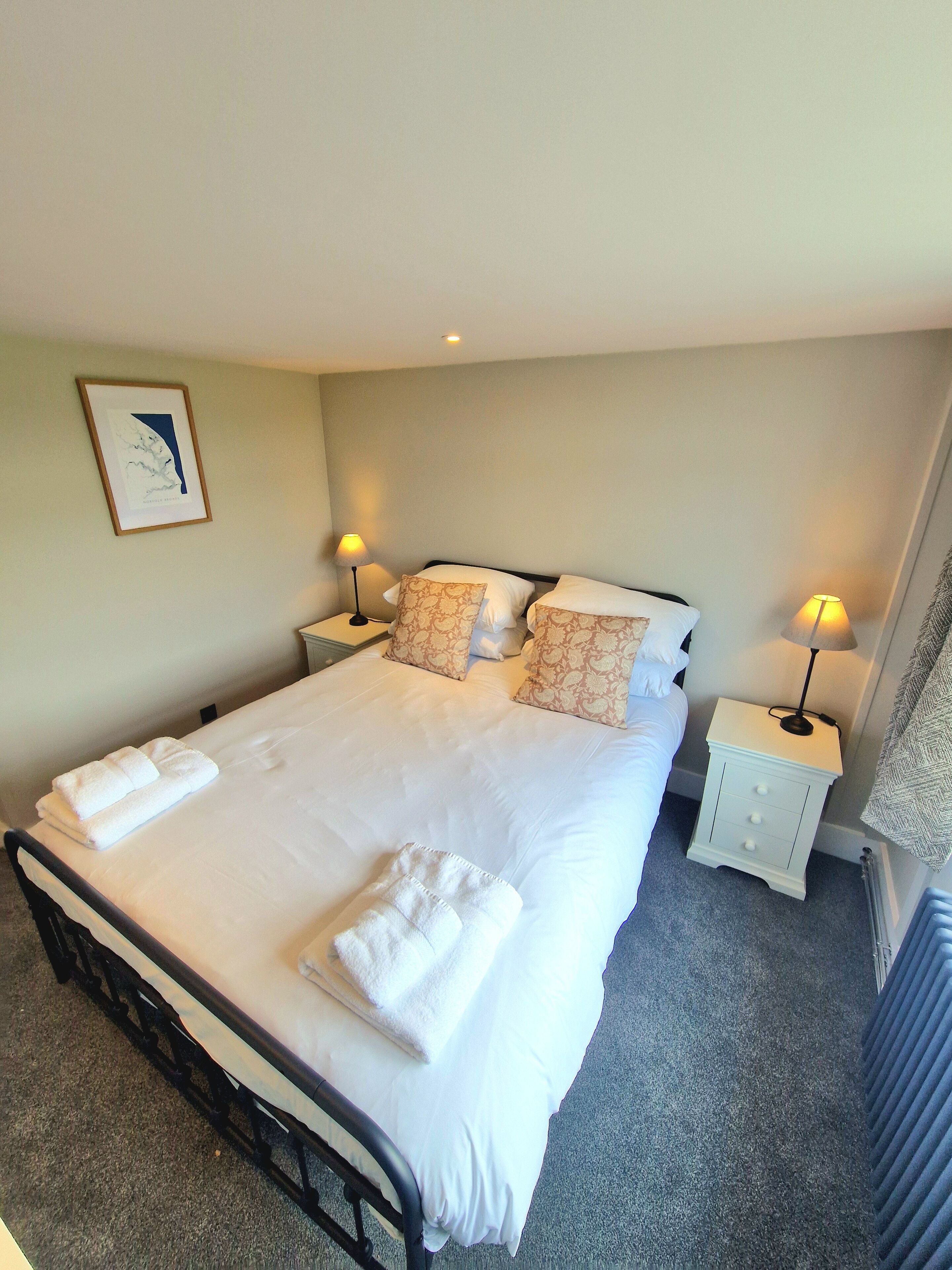 A neatly made bed with white linens and towels, two bedside lamps, a framed picture on the wall, and a radiator on the floor.
