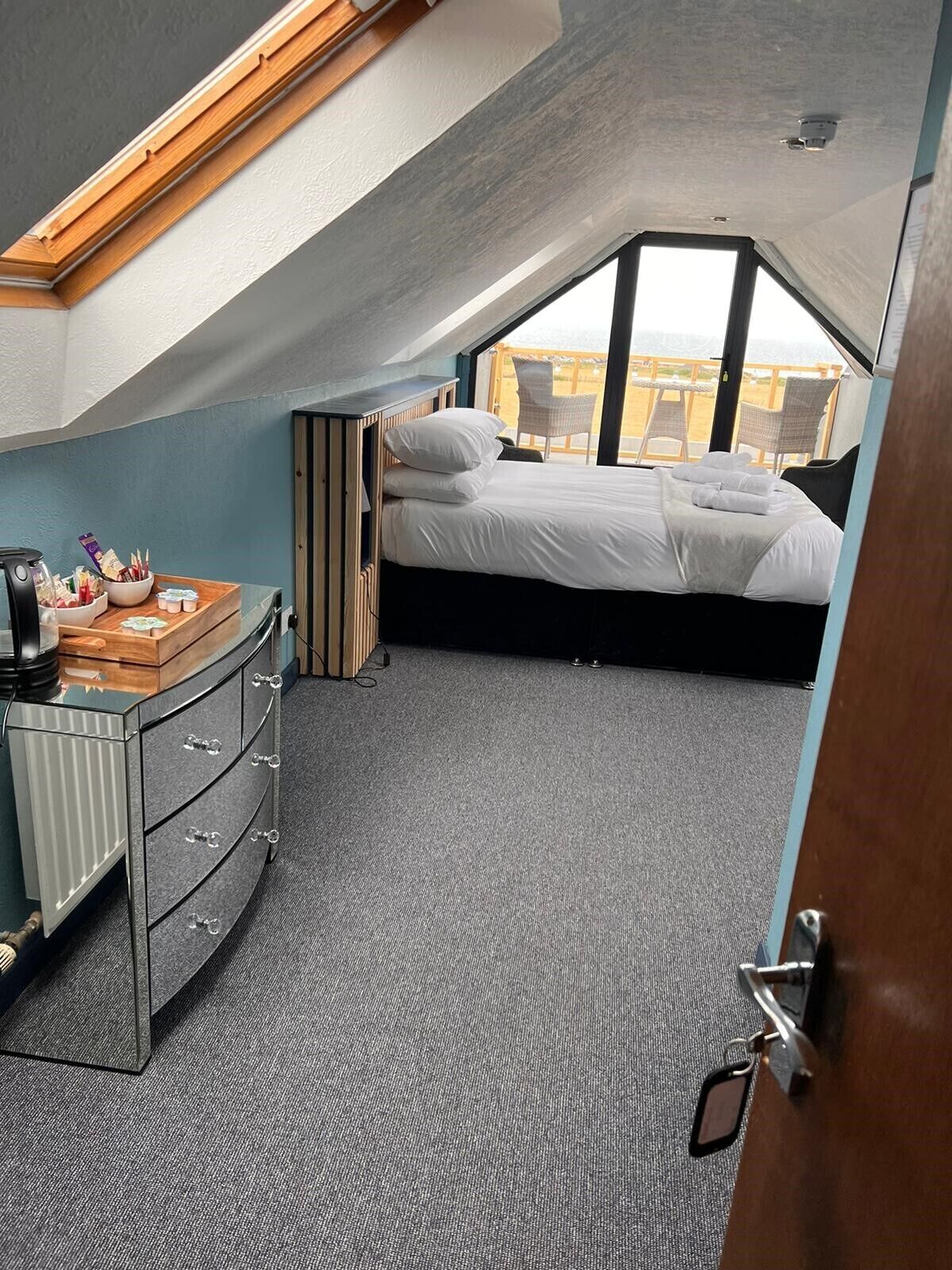 A bedroom with a bed, a dresser, a small table with toiletries, and a view of the beach through a window.