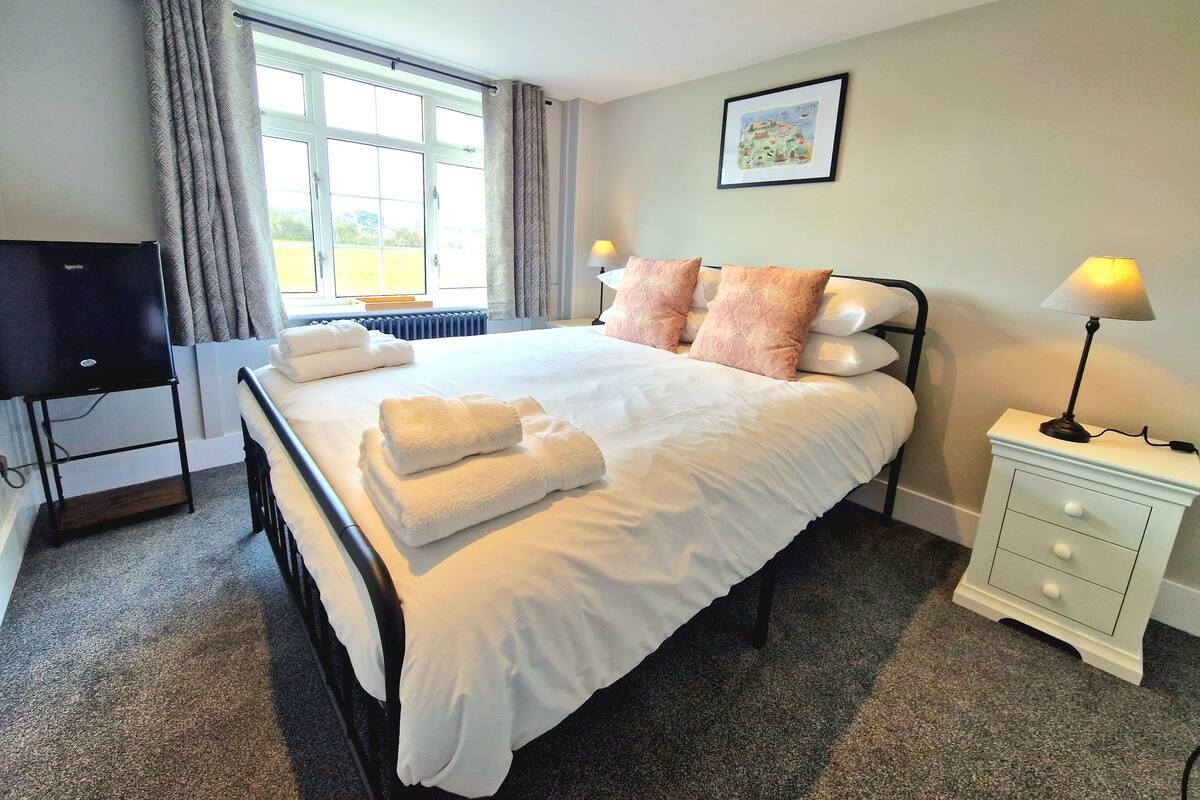 A neatly made bed with pillows, a bedside lamp, a TV on a stand, and a window with curtains.