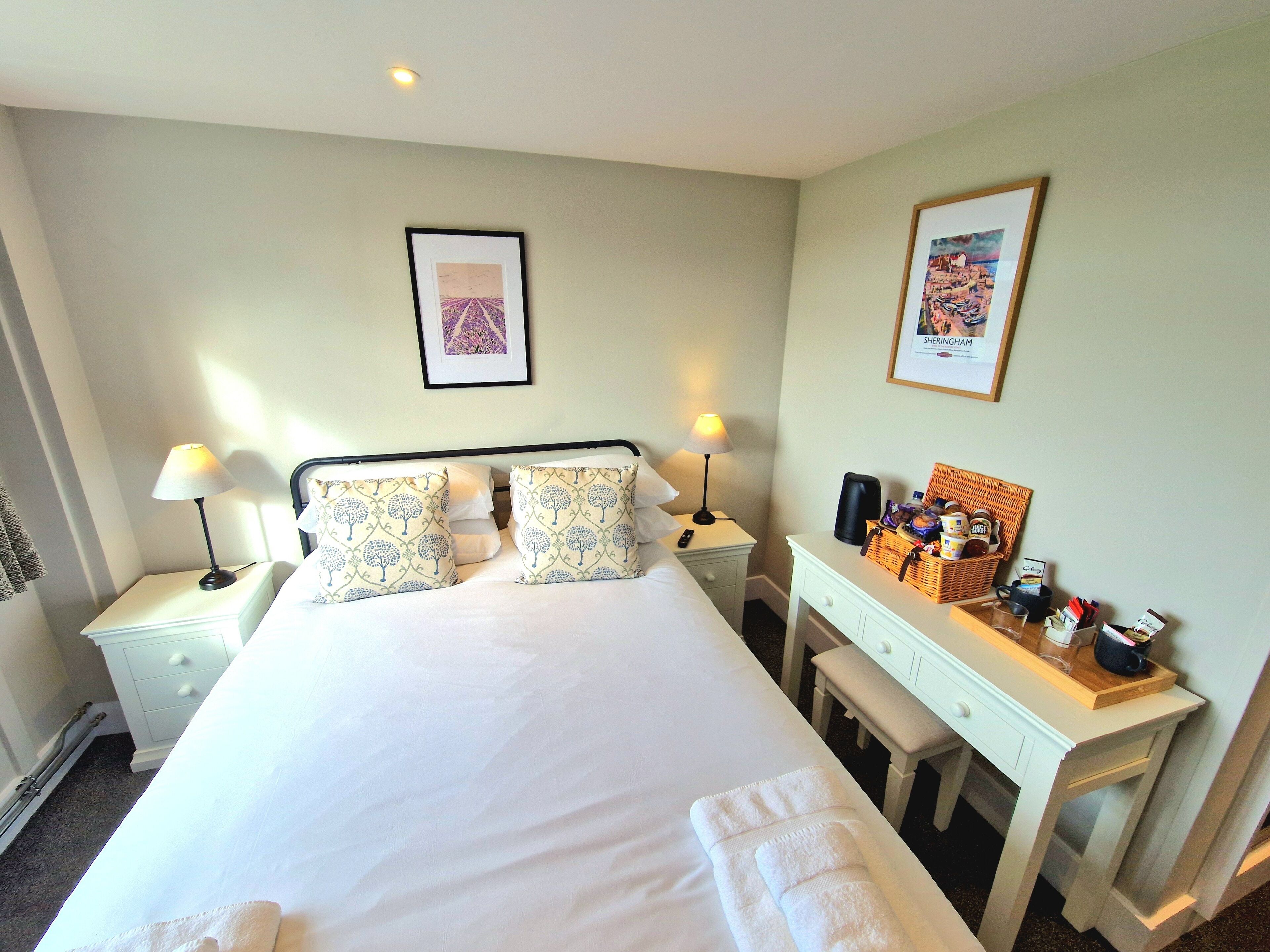 A neatly arranged hotel room with a large bed, two bedside tables with lamps, a small desk with a basket of toiletries, and two framed pictures on the wall.