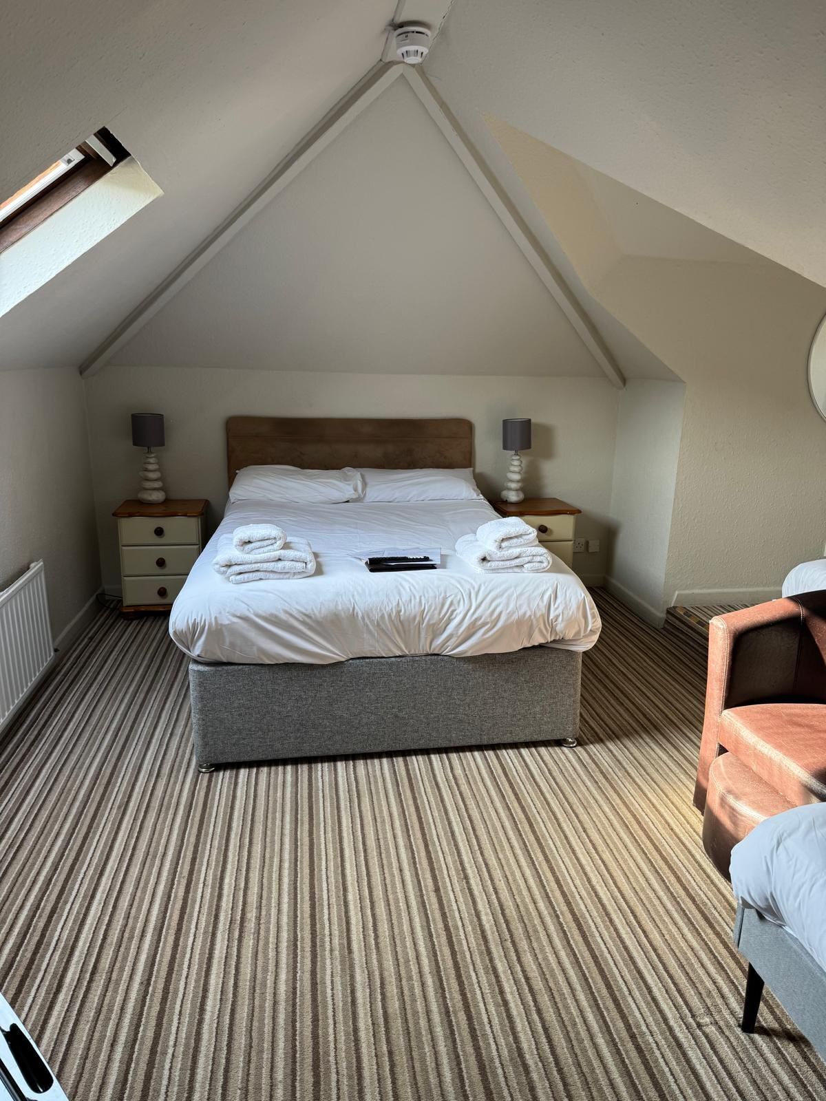 Ein Schlafzimmer mit einem großen Bett, zwei Nachttischen mit Lampen, einem Sessel und einer schrägen Decke.